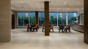 A spacious indoor area with multiple tables and chairs, large windows with a view of greenery, and a modern ceiling design. - Perch Service Apartments DLF Cyber City (Gurugram)