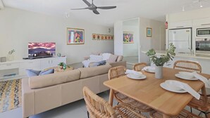 Ein modernes Wohnzimmer mit einer Couch, einem Esstisch und Stühlen. Ein Fernseher steht auf einem Schrank, und die Wände sind mit Kunstwerken geschmückt.