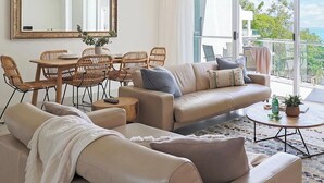 Ein modernes Wohnzimmer mit einem beigen Sofa, einem Esstisch mit Korbstühlen, einem Deckenventilator und einem großen Fenster mit Blick ins Freie.