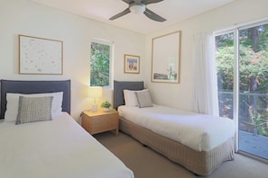 Two single beds with grey headboards, a nightstand, and a table with a lamp, surrounded by framed artworks and a large window with a view of the outdoors.