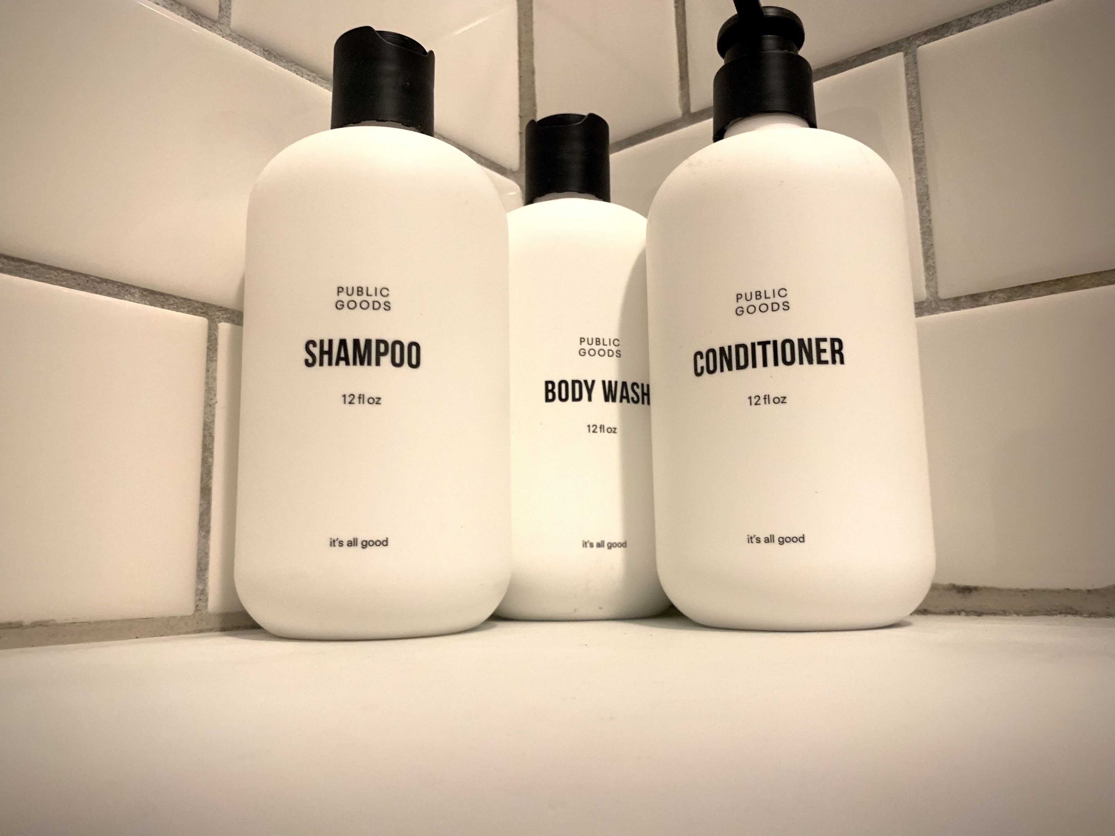 Três frascos de produtos da marca Public Goods: shampoo, gel de banho e condicionador, cada um com 354 ml.