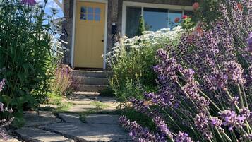 Une maison avec une porte jaune, une façade en pierre et un jardin où poussent différentes sortes de fleurs.