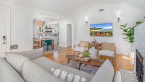 A spacious living room with a sofa, coffee table, and a fireplace. There is a dining area in the background with a view of the outdoors.