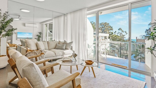 A modern living room with a large window, a sofa, two wooden chairs, a coffee table, and a view of the balcony and outside scenery. - Penthouse 20 - The Cove (Noosa)