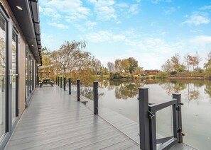 A wooden deck with a glass railing overlooking a calm body of water, trees, and a building with large windows.
