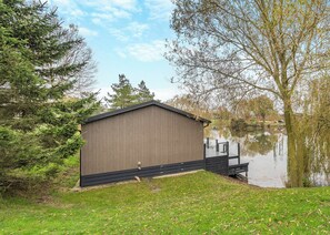 A small, single-story shed with a sloped roof, situated next to a body of water with a dock extending into the water.