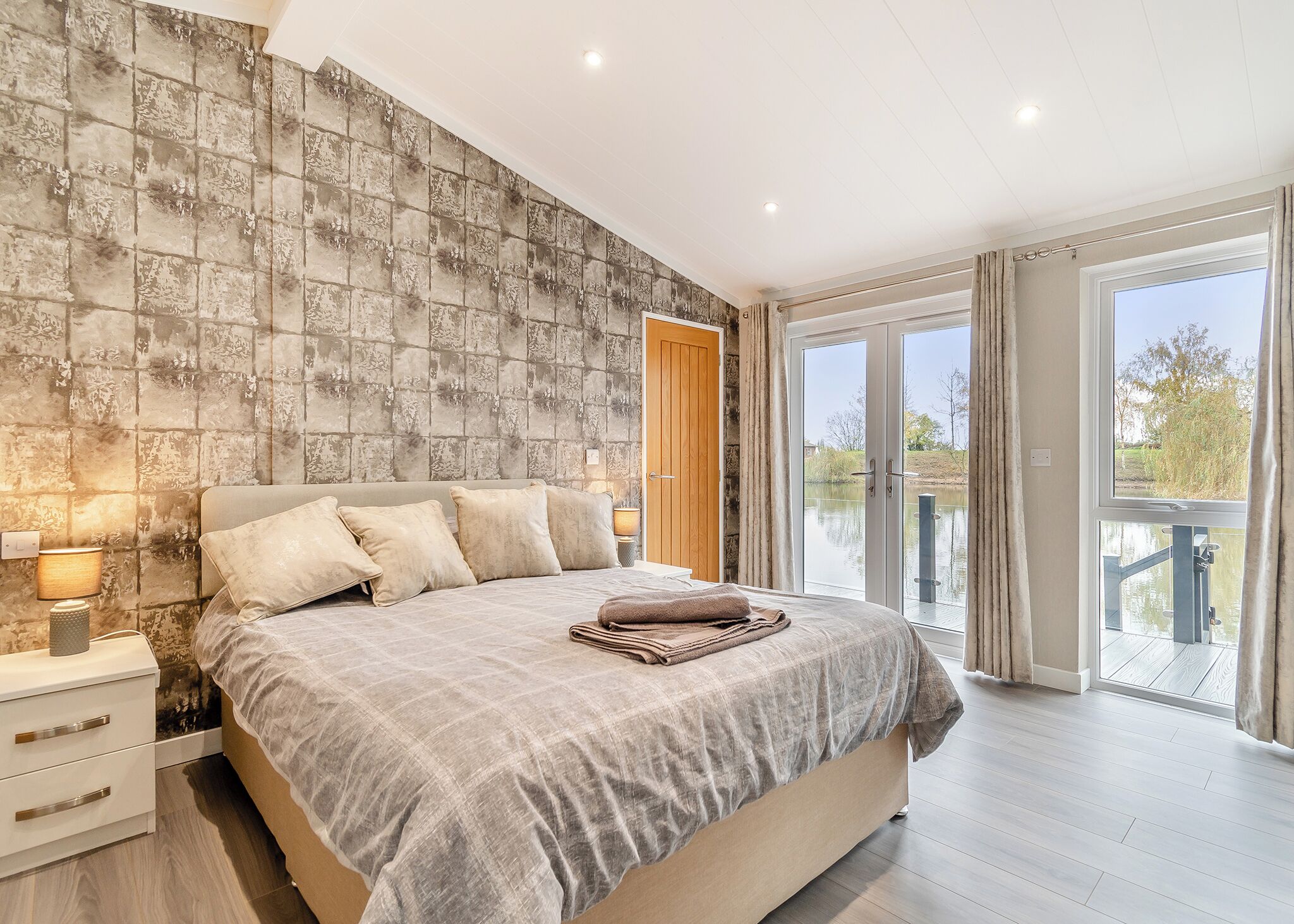 A bedroom with a large bed, a nightstand, a lamp, a wooden door, and a view of a body of water through a glass door.