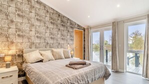 Une chambre à coucher avec un grand lit, une table de chevet, une lampe, une porte en bois et une vue sur une étendue d’eau, visible à travers une porte vitrée.