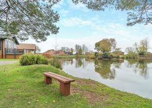 A lakeside view with a wooden bench, a building with a tiled roof, and a clear sky. - 1 bedroom accommodation in Swinethorpe, Nr Lincoln (Swinethorpe, Nr Lincoln)