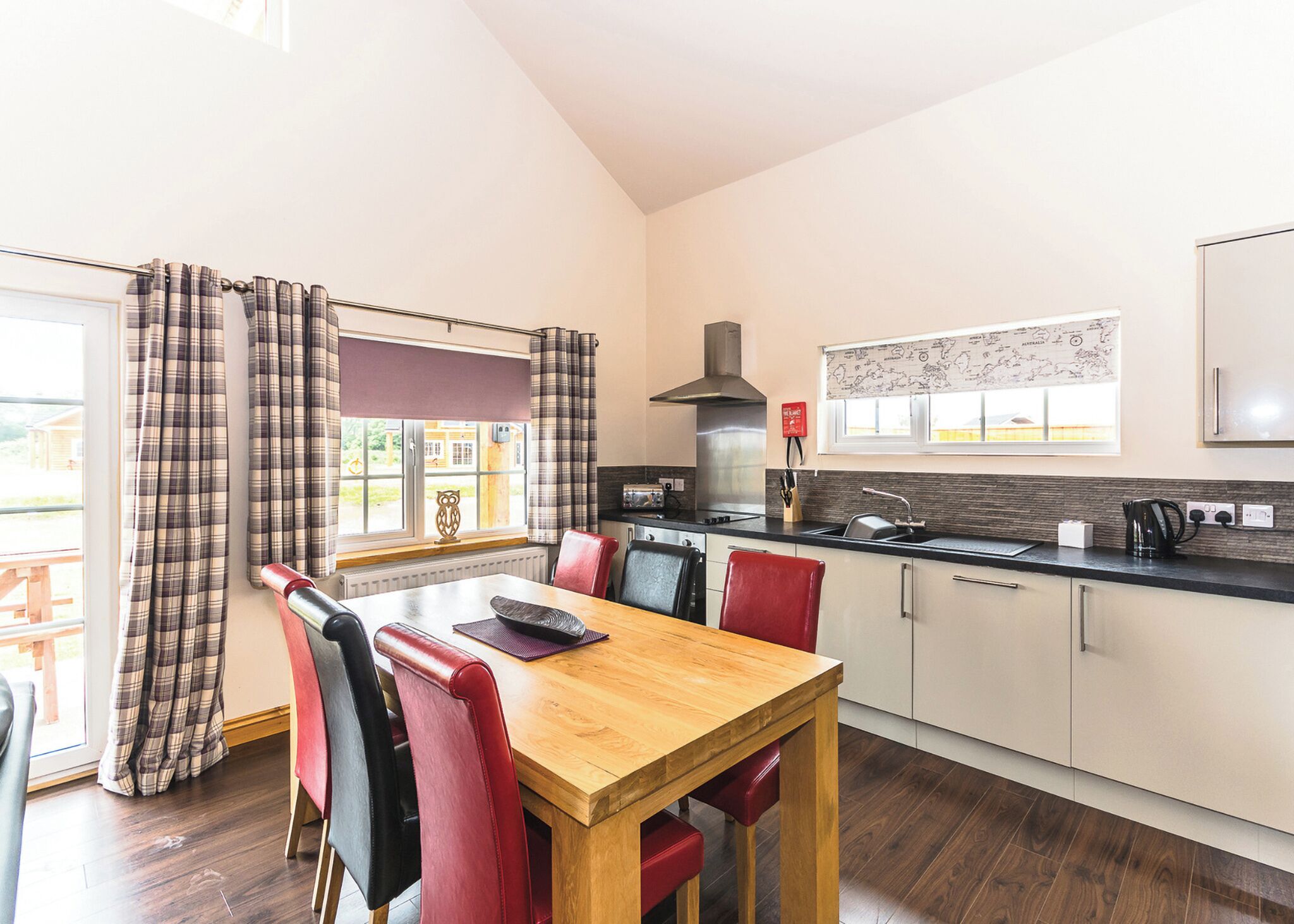 A modern kitchen with a dining area, featuring a wooden table, red and black chairs, a black stove, and a window with curtains.