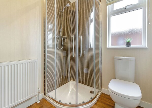 Et badeværelse med en brusekabine i glas, et hvidt toilet og et vindue med en plante.