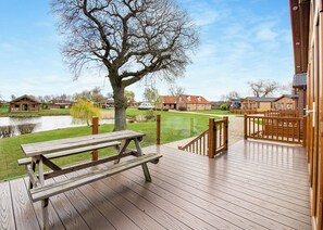Una terraza de madera con una mesa de picnic, un árbol y una vista de un lago y casas al fondo.