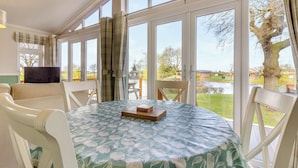 Un comedor con una mesa dispuesta para dos, ventanas amplias con cortinas, y vista a un lago y árboles.