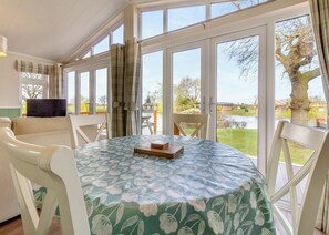 Un comedor con una mesa dispuesta para dos, ventanas amplias con cortinas, y vista a un lago y árboles.