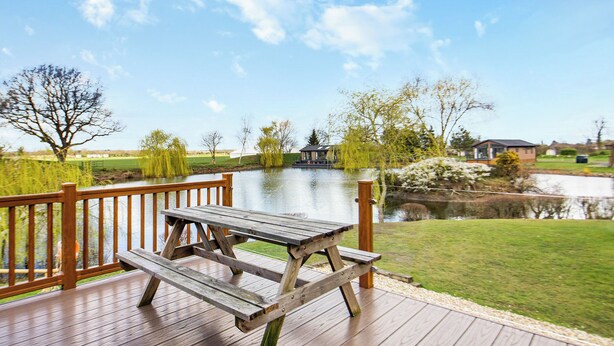 Una terraza de madera con una mesa de picnic que da a un lago, rodeada de árboles y un edificio pequeño a lo lejos.