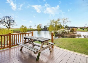 Une terrasse en bois avec une table de pique-nique, donnant sur un lac, entourée d’arbres et avec un petit bâtiment visible au loin.