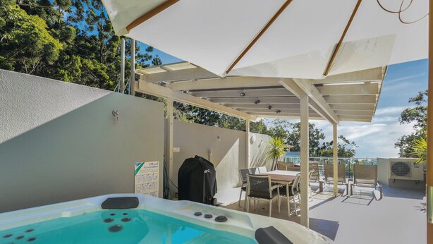 Ein Whirlpool unter einer überdachten Terrasse mit Tisch und Stühlen, umgeben von einer weißen Mauer und Bäumen.