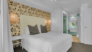 Ein Schlafzimmer mit einem großen Bett, einem Kopfteil mit floralem Wandpapier, zwei Nachttischlampen und Blick ins Badezimmer durch eine geöffnete Tür.