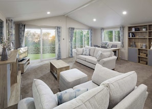 A spacious living room with a sofa set, a coffee table, and a TV. There are large windows with curtains, a bookshelf, and a dining area. - Platinum Lodge 4 Hot Tub (Skegness)