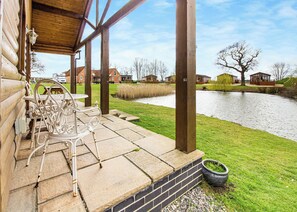 A wooden deck with outdoor seating, a brick wall, and a view of a river and houses in the background. - Traditional two bedroomed lodge with two double rooms. (Swinethorpe, Nr Lincoln)