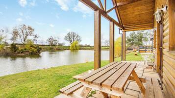 A wooden deck with picnic tables overlooking a calm lake, surrounded by trees and a grassy area.
