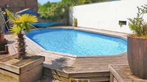 Una piscina con bordo in legno, immersa nel verde e circondata da un muro bianco.