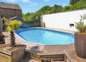 Une piscine dotée d’une terrasse en bois, entourée de verdure et bordée d’un mur blanc.