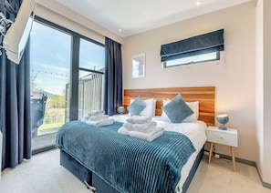 A modern bedroom with a large bed, a bedside table, a nightstand, a window with curtains, and a view of the outdoors.