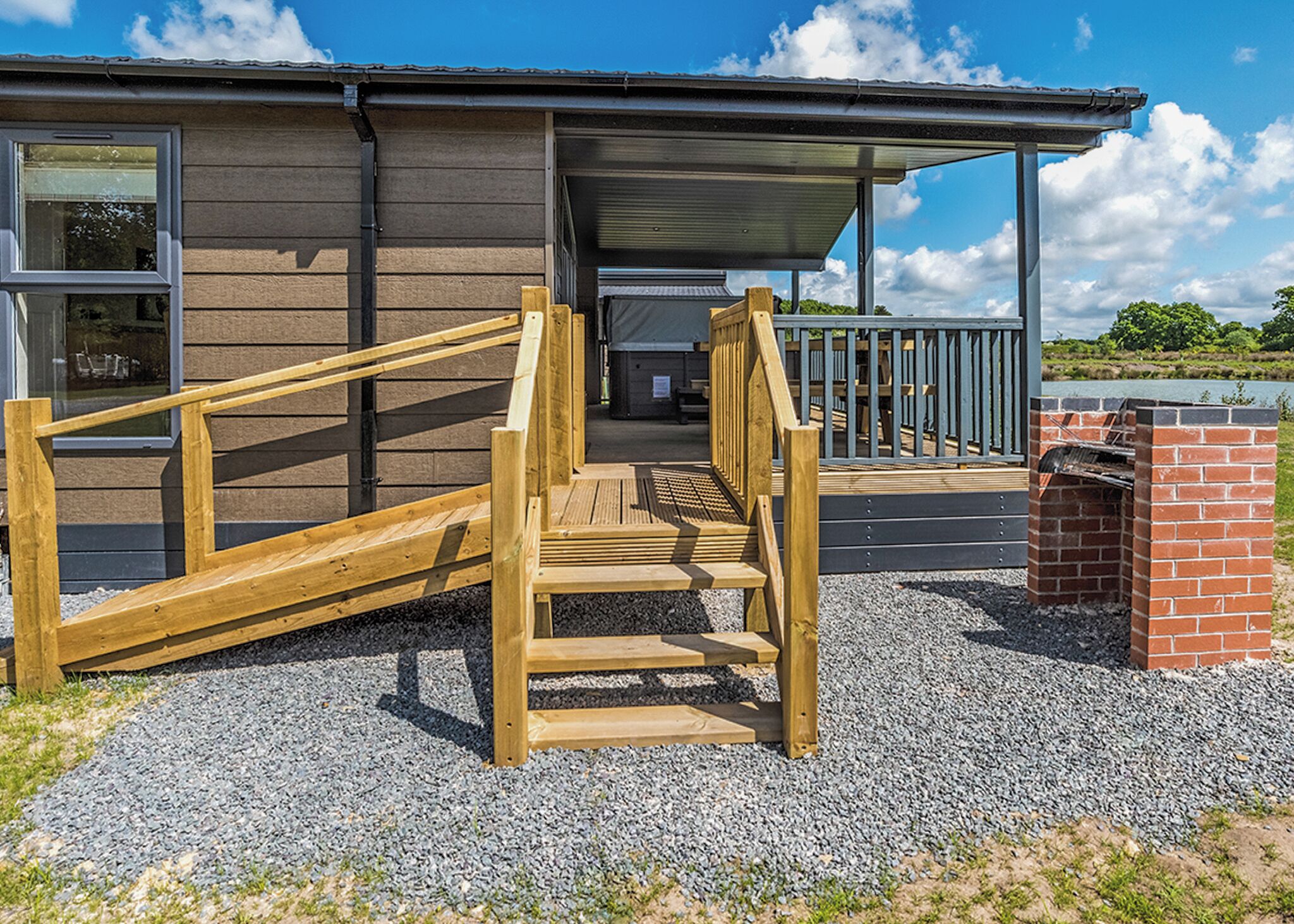 Un escalier en bois mène à une zone abritée, dotée d’une structure en briques, d’un sol en gravier et d’un bâtiment avec des fenêtres et un toit en métal.