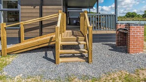 Un escalier en bois mène à une zone abritée, dotée d’une structure en briques, d’un sol en gravier et d’un bâtiment avec des fenêtres et un toit en métal.