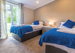 Ein Hotelzimmer mit zwei Betten, einem Nachttisch mit Lampe, einem Fenster mit Vorhängen und Blick auf Grünfläche.