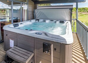A hot tub on an outdoor deck with a wooden bench, glass doors, and a view of the surrounding landscape.