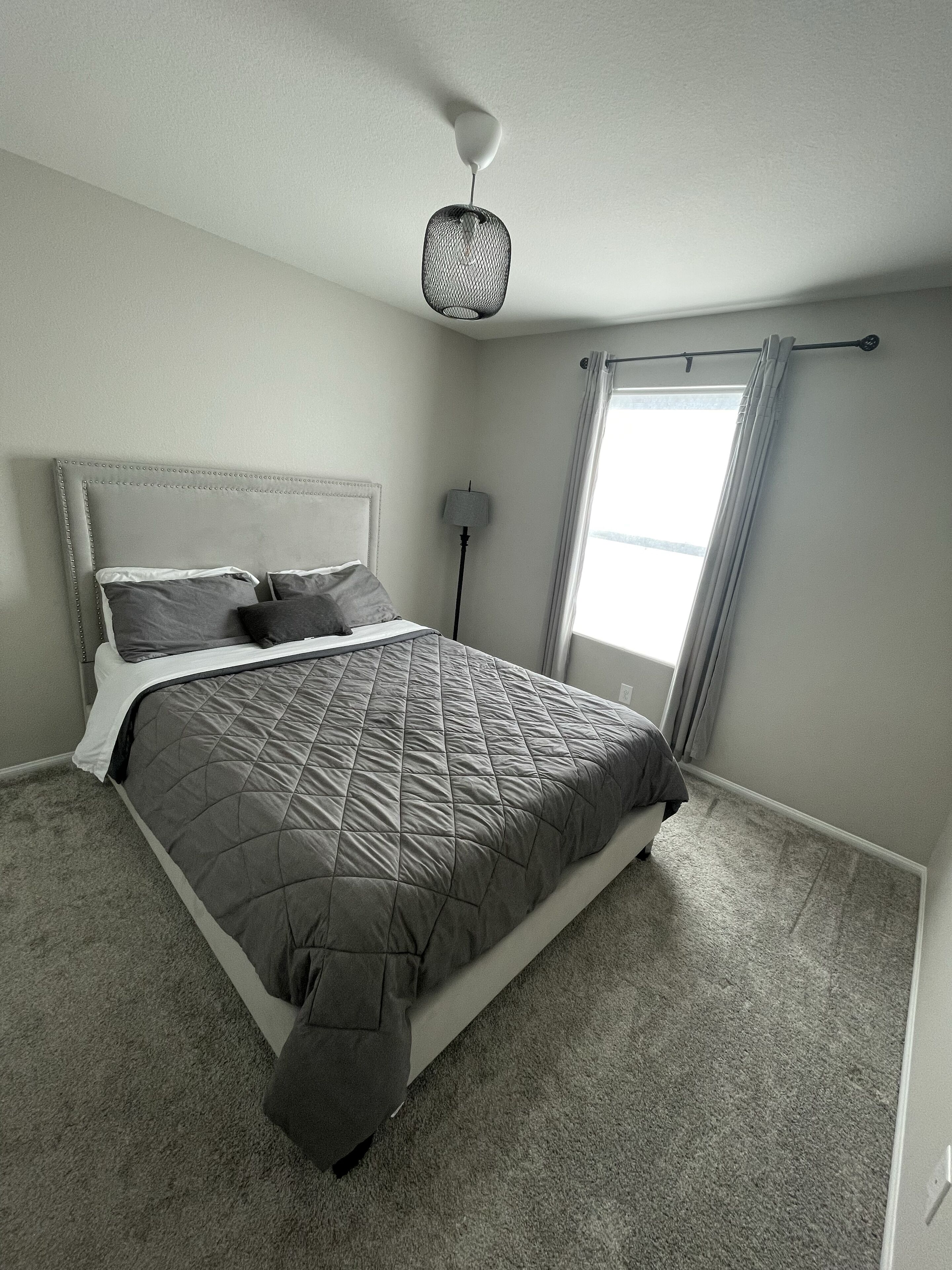 Un dormitorio con cama, lámpara de noche, ventana con cortinas y lámpara en el techo.