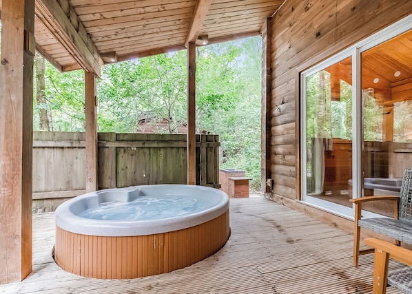A wooden cabin with a hot tub, a covered patio, and a view of the surrounding forest.