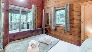 Una habitación tipo cabaña de madera con cama, dos ventanas con cortinas y un espejo.