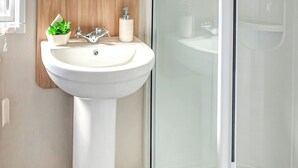 A bathroom with a white pedestal sink, a wooden panel above it, a small potted plant, a soap dispenser, and a glass shower enclosure. - 12ft-wide stylish and comfortable caravan with one double and one twin. (Chapel St Leonards)