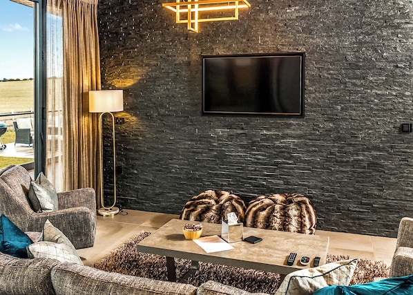 A modern living room with a stone wall, a flat-screen TV, a coffee table, and comfortable seating. - Four bed lodge with three king-size beds and one twin (king by arrangement). (Nottingham)