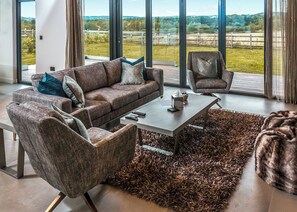 A modern living room with a sofa, armchair, and coffee table, large windows with a view of a green landscape. - Four bed lodge with three king-size beds and one twin (king by arrangement). (Nottingham)