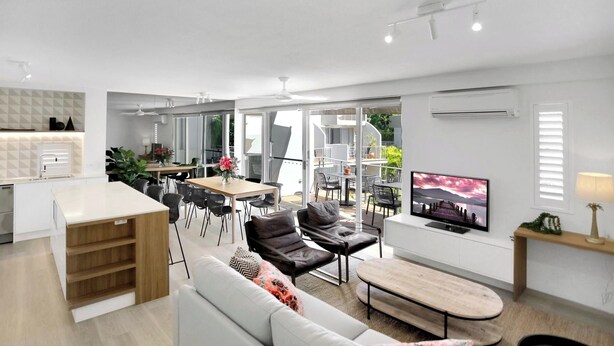 A modern kitchen with a dining area, a living room with a sofa and coffee table, and a TV on the wall. - Penthouse 22  - The Cove (Noosa Heads)