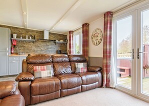 A living room with a brown leather sofa, a stone wall, a kitchen with white cabinets, and a large window with curtains.