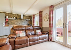 A living room with a brown leather sofa, a stone wall, a kitchen with white cabinets, and a large window with curtains.