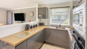 A modern kitchen with a built-in oven, a large window with blinds, a flat-screen TV, and a coffee maker. - Lakeview Premier Pet Friendly (Woodhall Spa)
