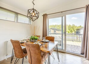 Ruokailutila, jossa on puinen pöytä, ruskeat tuolit, riippuvalaisin sekä näkymä ulkona olevalle terassille ja rakennuksille.