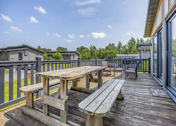 Une terrasse en bois avec des tables et des chaises pour pique-niquer, donnant sur une pelouse, avec des bâtiments visibles à l’arrière-plan.