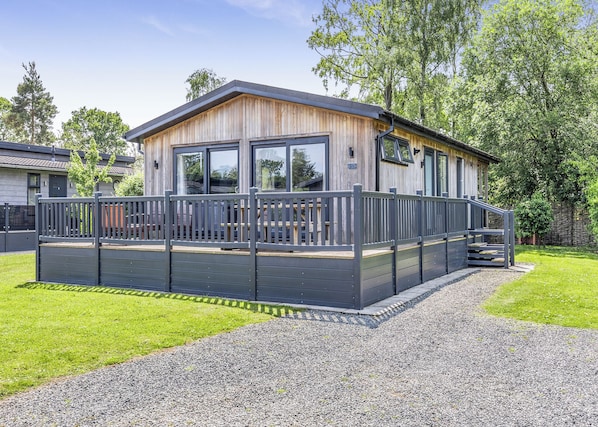 A modern wooden cabin with a deck, large windows, and a gravel driveway.