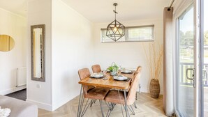 A dining area with a wooden table set for four, a geometric pendant light, and a view through a glass door. - Lakeview Premier Pet Friendly (Woodhall Spa)