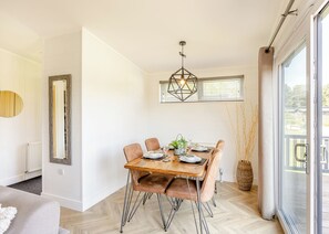 Un coin repas avec une table en bois dressée pour quatre personnes, un luminaire suspendu aux formes géométriques et une vue à travers une porte vitrée.