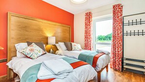 Een hotelkamer met twee bedden, een houten hoofdeinde, een nachtkastje met een lamp, een raam met gordijnen en een kapstok.