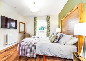 A hotel room with a bed, a TV, a chair, a window with curtains, a radiator, and a wooden floor. - 3 bedroom accommodation in Flaxton (Flaxton)