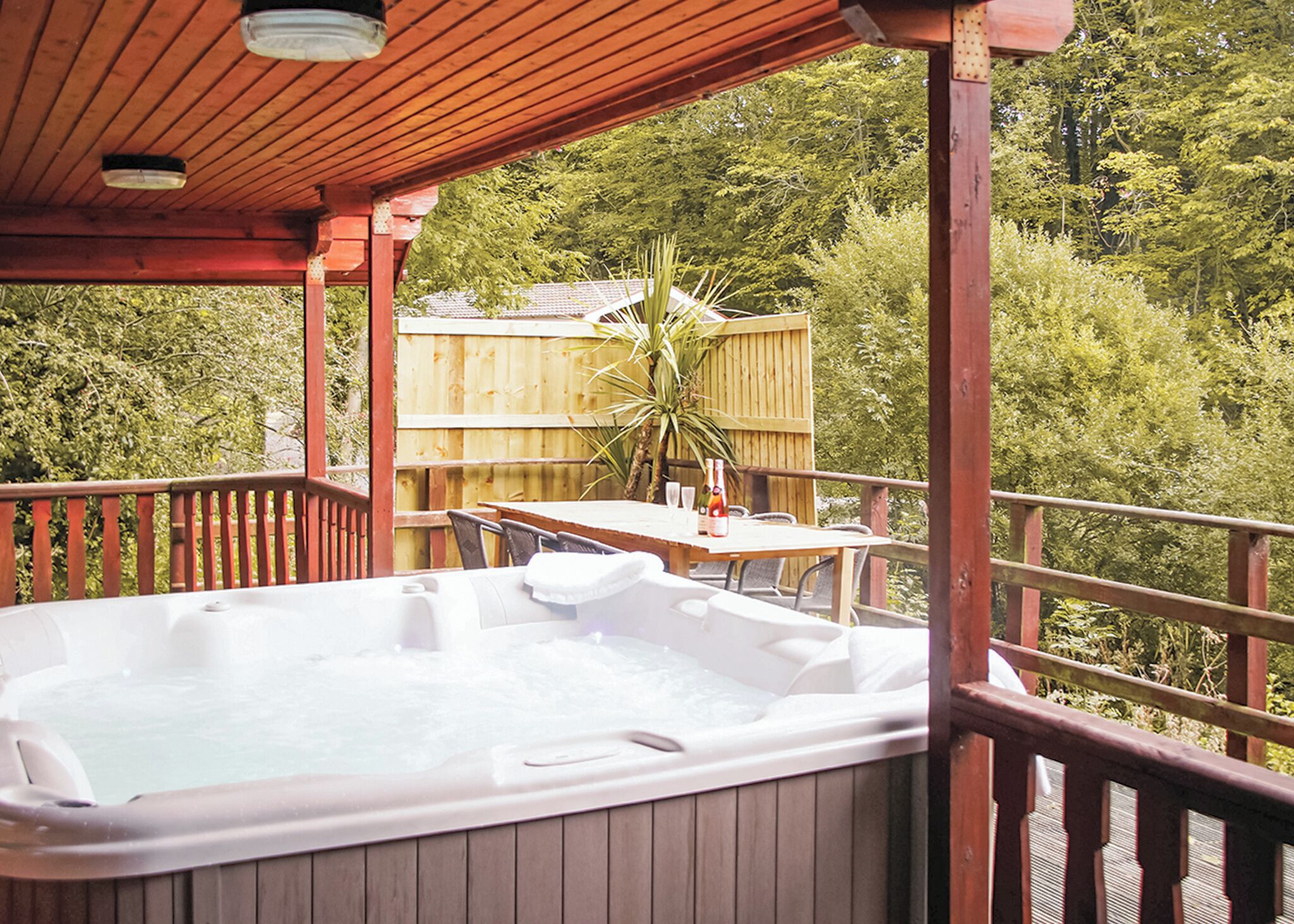Un bain à remous sur une terrasse en bois, avec une table et des chaises, entouré d’une clôture en bois et d’arbres.